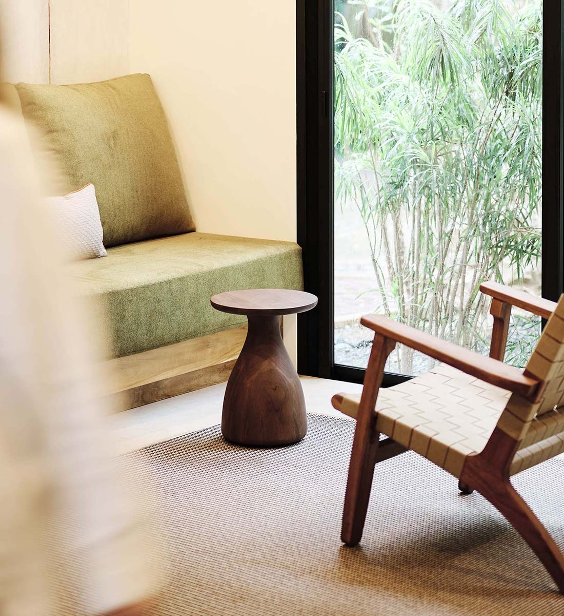 A styled interior corner featuring a low upholstered bench with a large olive-green cushion, a small round wooden side table, and a woven-back wooden chair with a warm wood frame, set on a textured rug beside a large window overlooking bamboo.