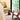A styled interior corner featuring a low upholstered bench with a large olive-green cushion, a small round wooden side table, and a woven-back wooden chair with a warm wood frame, set on a textured rug beside a large window overlooking bamboo.