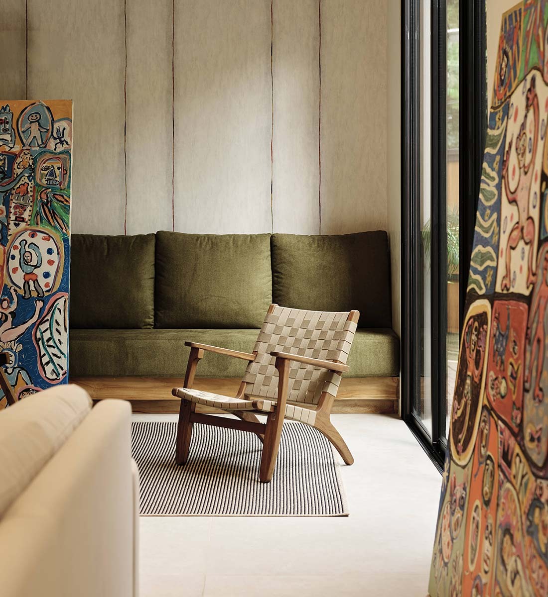 A mid-century modern wooden armchair with a woven seat and back is placed on a striped rug in a styled living room. Behind the chair is an olive green velvet sofa against a beige paneled wall, with colorful abstract art panels visible on both sides.